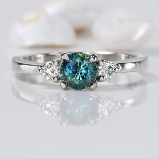 Platinum trilogy engagement ring with teal sapphire and two round diamonds on reflective surface.