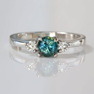 Handmade platinum trilogy engagement ring with teal sapphire and two round diamonds on reflective surface.