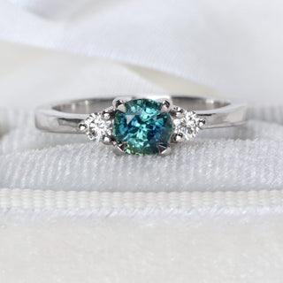 Close-up of handmade platinum trilogy engagement ring featuring teal sapphire and round diamonds.