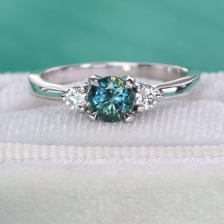 Close-up of teal sapphire and diamond trilogy engagement ring in handmade platinum on green background.