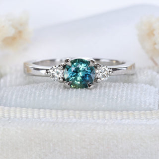 Platinum trilogy engagement ring with teal sapphire and diamonds photographed on pale fabric.