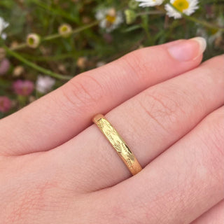 Handmade sustainable solid 18ct gold 3mm court wedding ring with hand engraved lavender flowers around the band.  Shown being worn on the hand with daisies in the background