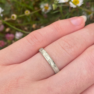 Platinum 3mm court wedding ring with hand engraved lavender flowers around the band. Shown being worn on the hand with daisies in the background