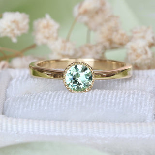 Close-up of handmade 18ct gold solitaire engagement ring with green sapphire in a milgrain bezel setting.