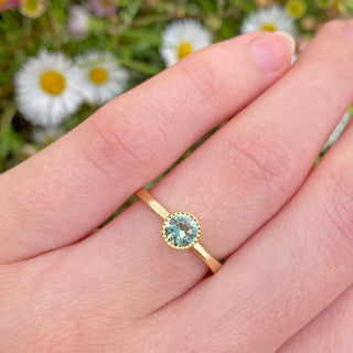 Handmade 18ct gold solitaire engagement ring bezel set with a unheated 0.70 carat green sapphire, with pretty milgrain detail around the stone. Displayed worn on the hand with daisies in the background