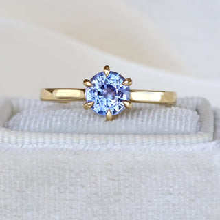 Close-up of lavender blue sapphire solitaire engagement ring in 18ct gold on pale fabric.