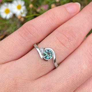 Handmade platinum wave engagement ring set with a fair trade natural ocean teal sapphire and white diamond accent. Show here being worn on the hand with daisies in the backdrop