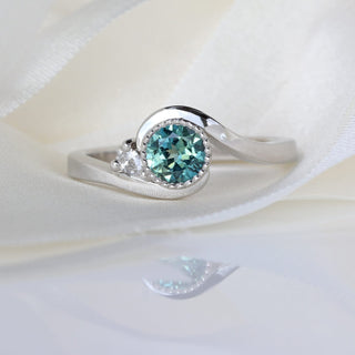 Platinum teal Parti sapphire engagement ring with wave-shaped band photographed on pale fabric.