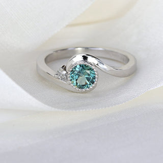 Close-up of platinum wave engagement ring with teal Parti sapphire bezel and pear diamond accent.