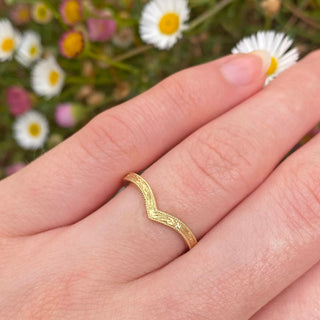 Handmade sustainable solid 18ct gold wishbone wedding band with floral engraved orange blossom detail, framed by a milgrain border. Displayed in the photo on the hand with daisies in the background