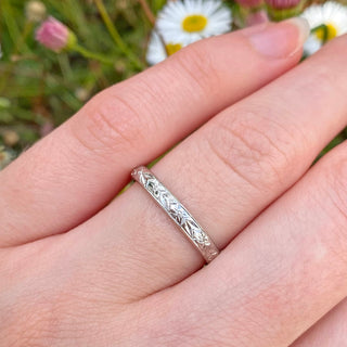 Hand engraved 2.5mm wide court shape platinum wedding ring with a hand engraved orange blossom and leaves pattern around the band. Being worn on the hand with daisies in the background 