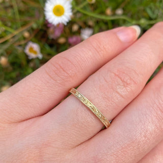 Solid 18 gold 2mm wide wedding band with hand engraved orange blossom and leaves around the band, framed in a milgrain border. Displayed worn on the hand with flowers in the distance