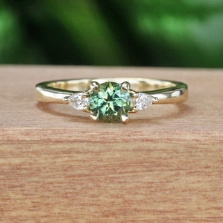 18ct gold three stone engagement ring with a green sapphire and pear cut diamonds, on a wooden surface