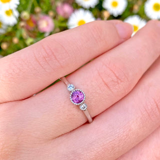 Petite platinum trilogy ring set with a 4mm pink sapphire and natural white diamonds.  Displayed on the hand with daisy backdrop