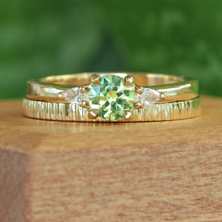 Close up of green sapphire and pear cut diamond trilogy engagement ring and slim tree bark textured wedding ring on a wooden box with blurred green background