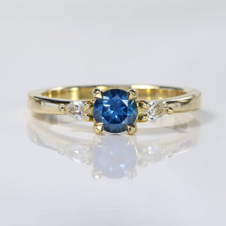 Handmade 18ct gold blue sapphire trilogy engagement ring with pear cut diamonds on reflective surface.