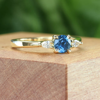 Side angle of fairtrade blue sapphire and diamond trilogy engagement ring on a wooden box with green backdrop.