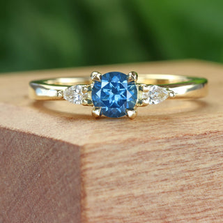Handmade 18ct gold trilogy engagement ring with blue sapphire and pear diamonds on wooden surface.