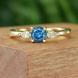 Handmade 18ct gold trilogy engagement ring with blue sapphire and pear diamonds on wooden surface.