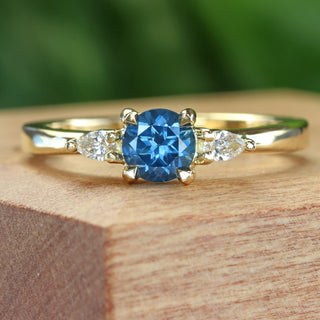Handmade 18ct gold trilogy engagement ring with blue sapphire and pear diamonds on wooden surface.
