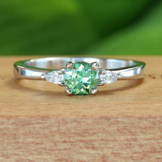 Handmade platinum trilogy engagement ring with green sapphire and pear cut diamonds on wooden ring box.