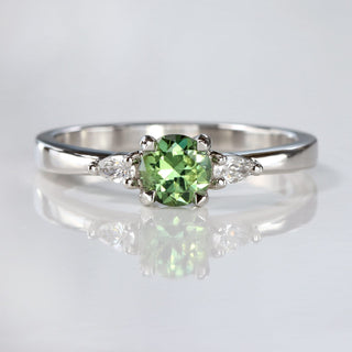 Handmade platinum trilogy engagement ring with green sapphire and pear cut diamonds on reflective surface.