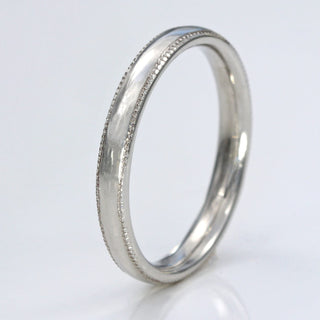 Slim platinum court wedding band with hand engraved milgrain border, resting on a white surface