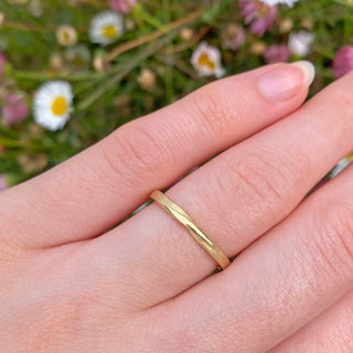Handmade solid 18ct gold wedding ring with a twist in the band at the front of the ring. The band has Lilia's signature spun silk texture applied. Displayed on the hand with daisies in the background