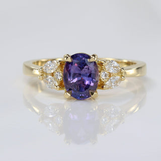 Handmade 18ct gold oval purple sapphire engagement ring with diamond accents on reflective surface.