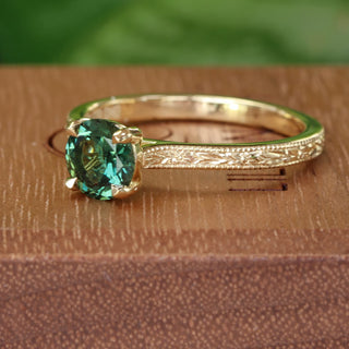 Angled view of a green sapphire solitaire engagement ring, handmade from sustainable 18ct gold. Hand engraved floral pattern on the band