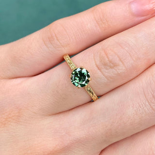 18ct gold solitaire engagement ring with engraved orange blossom detail, set with a green sapphire being worn in the hand