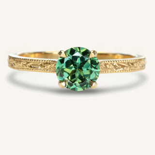 Handmade 18ct gold solitaire engagement ring set with a fair trade green sapphire. The 2mm wide band has hand engraved floral orange blossom and leaves pattern