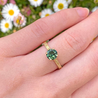 18ct gold solitaire engagement ring set with a fair trade green sapphire. The band has been hand engraved with a floral orange blossom and leaves pattern. Displayed being worn on the hand with daisy backdrop