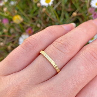 Handmade 18ct gold slim 2mm wide wedding band with a tree bark hammered texture applied. Shown being worn on the hand with daisies in the background.