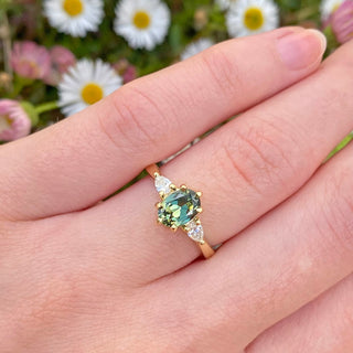 Veryan 18ct gold trilogy engagement ring set with a fair trade oval Parti sapphire and pear cut natural white diamonds. Featured being worn on the hand with daisy backdrop