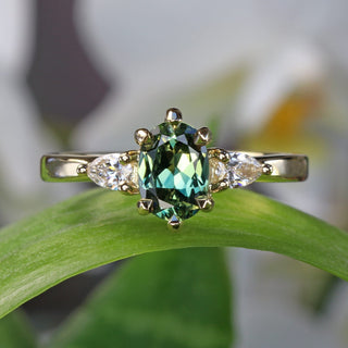 18ct gold oval fair trade green sapphire and pear cut diamond trilogy engagement ring on a leaf with blurred white flowers