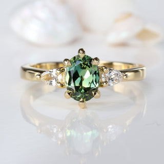 18ct gold oval fair trade green sapphire and pear cut diamond trilogy engagement ring on a white surface