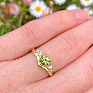 Zennor handmade 18ct gold wishbone engagement ring set with a fair trade green sapphire and natural white diamonds, with milgrain detail around the settings. Being worn on the finger with daisy background