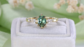 18ct gold three stone engagement ring with oval green sapphire and side diamonds on a white velvet box with green background