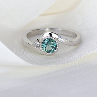 Close-up of platinum wave engagement ring with teal Parti sapphire bezel and pear diamond accent.