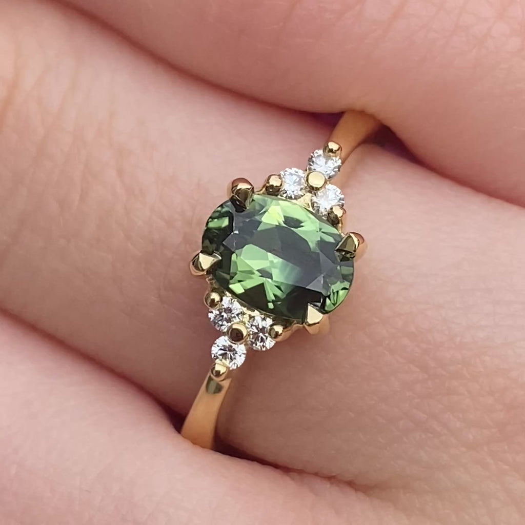 Handmade Oval Parti sapphire and diamond cluster engagement ring in 18ct yellow gold shown on hand.