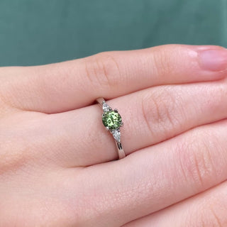 Platinum trilogy engagement ring with green sapphire and diamonds shown on hand.