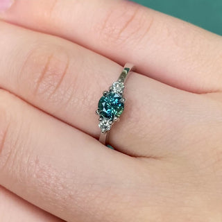 Handmade platinum teal sapphire trilogy engagement ring shown worn on hand.
