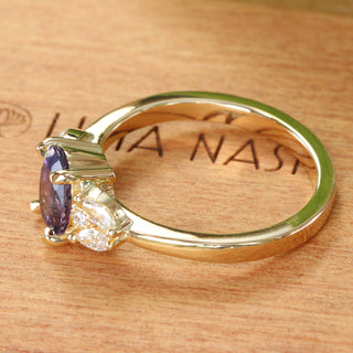Side profile of purple sapphire oval and diamond cluster engagement ring in 18ct gold. Placed on branded wooden ring box.