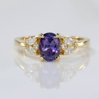 Handmade 18ct gold oval purple sapphire engagement ring with diamond accents on reflective surface.