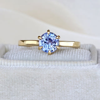 Close-up of lavender blue sapphire solitaire engagement ring in 18ct gold on pale fabric.