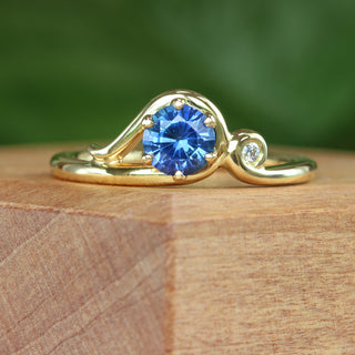 Blue sapphire engagement ring with Art Nouveau curl design photographed on wooden surface.