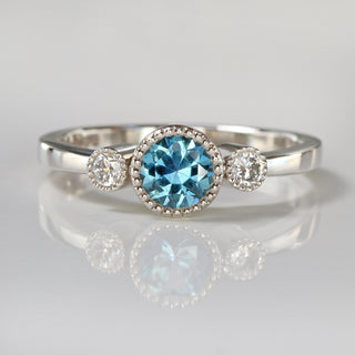 Handmade platinum trilogy engagement ring with teal blue Montana sapphire and Canadian diamonds on reflective surface.