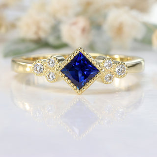 Princess cut blue sapphire and diamond cluster ring on white floral backdrop.