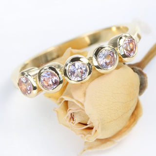 A gold ring with five peachy pink sapphires set in bezel-like settings, displayed on a white surface next to a dried flower.